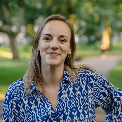 jonge vrouw met laflang donkerblond haar in blauwe bloes met dessin tegen een achtergrond van een parkachtige omgeving